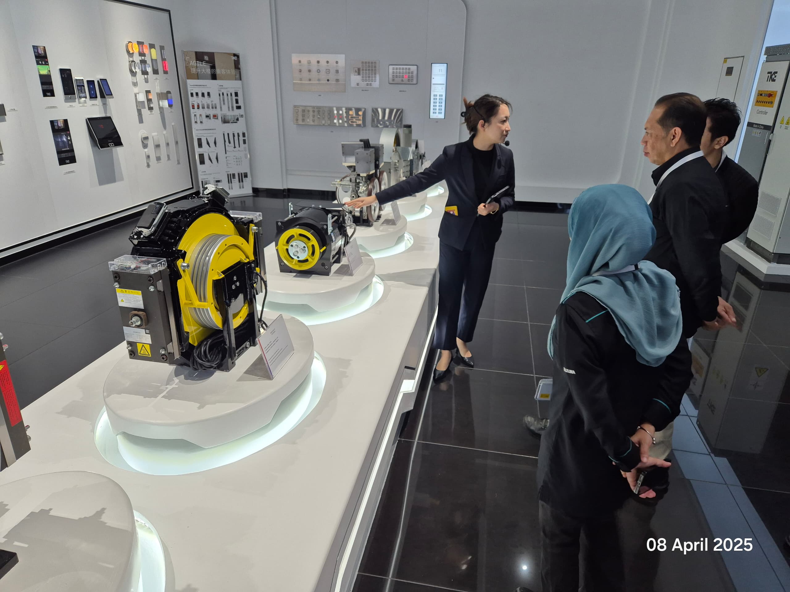 Gearless traction machines displayed on lit pedestals while a TKE host explains them to the visitors